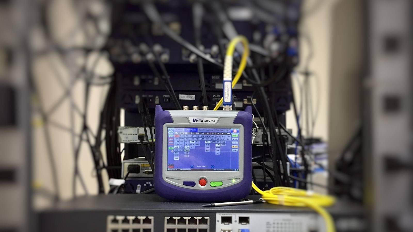 Analisador de rede óptica conectado por cordão monomodo a um switch, medindo alarmes em um rack